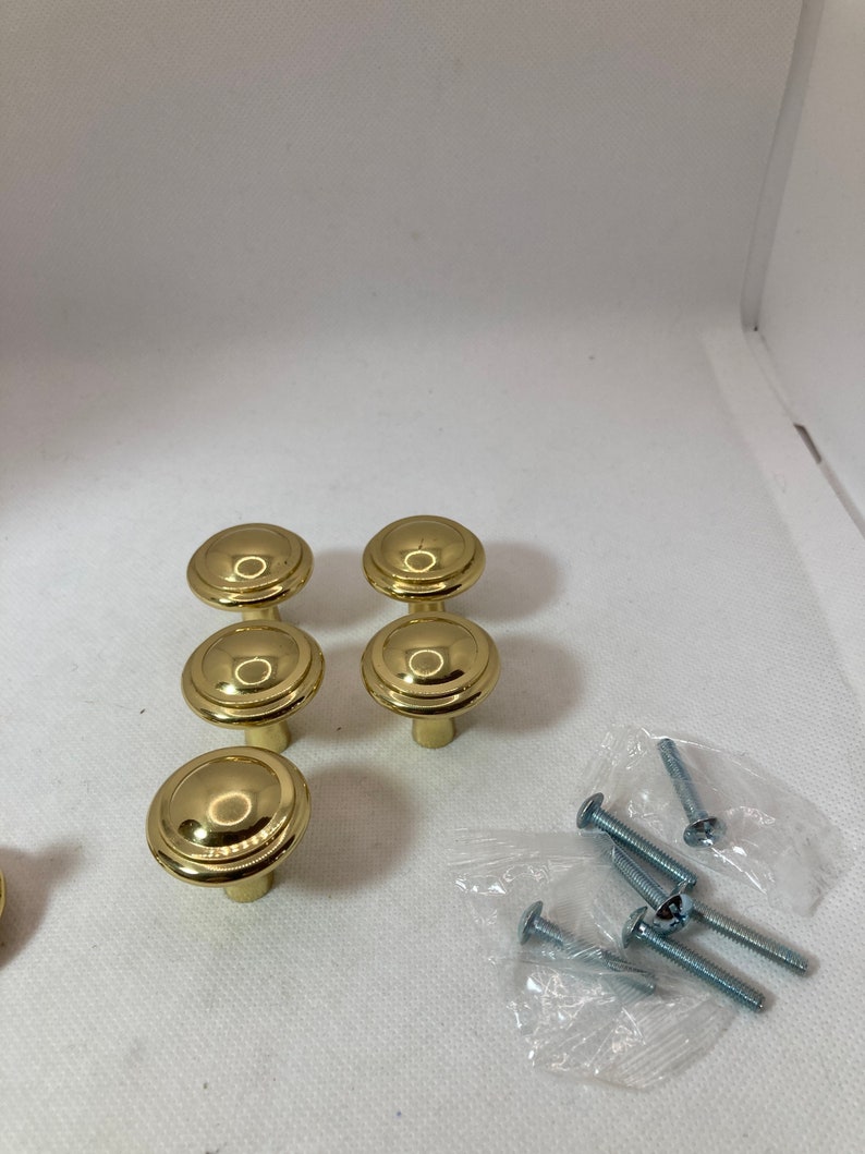 以下が含まれることがあります： 丸みを帯びた段付きデザインの、金色のキャビネットノブ6個セット。ノブは3つずつ2列に配置され、近くには小さなネジのパックが見えます。ノブは光沢のある反射面を持っています。