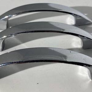 May include: Three chrome-finished cabinet handles. Each handle has a curved, arched design with a smooth, reflective surface. The handles are made of metal and are suitable for drawers or cabinets.