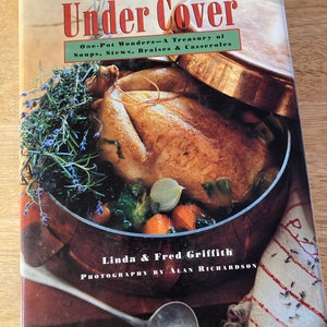 May include: A cookbook titled "Cooking Under Cover" featuring a roasted chicken in a pot. The cover includes text about one-pot wonders, soups, stews, braises, and casseroles. The book is by Linda & Fred Griffith, with photography by Alan Richardson.