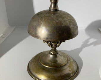 May include: Antique silver-toned bell with a dome-shaped top and a round base. The bell has a small, decorative handle and a spring mechanism. The base has a decorative beaded edge. The bell is likely made of metal and has a vintage appearance.