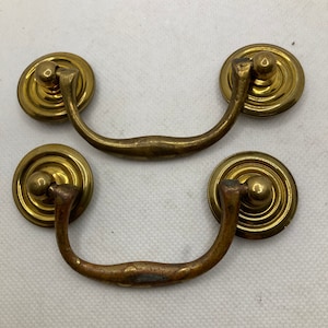 May include: Two antique brass drawer pulls. Each pull has a circular backplate with a raised, concentric ring design. A curved handle connects to the backplate. The brass has a warm, aged patina, with some areas showing darker oxidation.