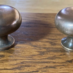 May include: Two antique bronze cabinet knobs with a rounded top and a short, flared base. The knobs are set against a wooden surface, showcasing their metallic finish and classic design. These knobs are ideal for adding a vintage touch to furniture.
