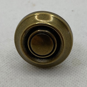 May include: A close-up of a bronze-colored cabinet knob. The knob has a circular design with concentric rings and a raised center. The finish has a slightly aged appearance, suggesting a vintage or antique style. The knob is likely used for furniture or cabinetry.