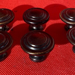 May include: Six bronze-colored cabinet knobs with a layered, circular design. The knobs have a dark, metallic finish with a lighter bronze edge. Screws are visible in the background. The knobs are on a red surface.