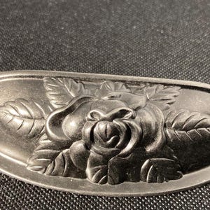 May include: A dark gray cabinet pull with a raised rose and leaf design. The hardware is oval-shaped with a detailed floral pattern. Two screws are visible in the background.