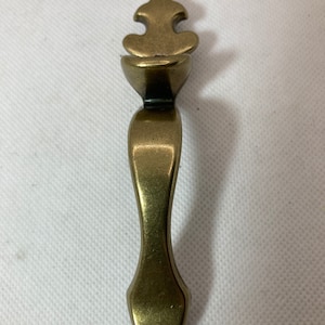May include: A bronze-colored cabinet pull with an ornate, stylized top and a curved handle. The pull has a vintage aesthetic, suitable for furniture restoration or adding a classic touch to cabinetry. The overall length is approximately 5 inches.