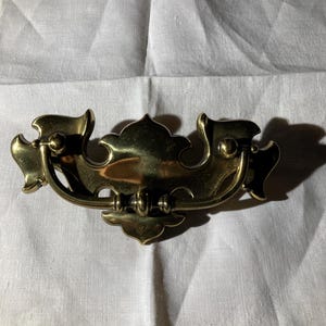 May include: A brass drawer pull with an ornate, symmetrical design. The pull features a central handle with decorative flourishes on either side. The brass has a polished finish, reflecting light. The overall shape is curved and elegant, suitable for furniture.