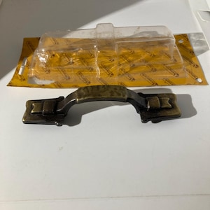 May include: A bronze-toned cabinet pull handle with a curved grip and rectangular end pieces. The handle is in its original packaging, a clear plastic blister pack on a yellow and white card.