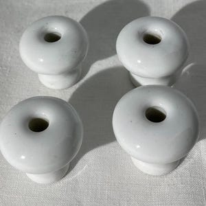 May include: Four white ceramic cabinet knobs with a donut-like shape and a central hole. The knobs are round with a small base and are arranged on a white surface. The knobs are likely for furniture or decorative use.