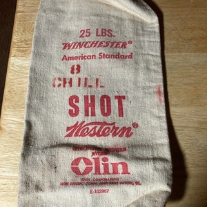 May include: A tan canvas bag with red lettering. The text reads "25 LBS. WINCHESTER American Standard, CHILL SHOT, Western, Olin Corporation". The bag is slightly crumpled and appears to be vintage.