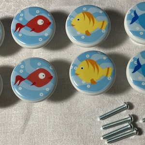 May include: Set of eight ceramic cabinet knobs with colorful fish designs. Each knob features a different fish in shades of green, red, yellow, and blue, set against a light blue background with white bubbles. Includes mounting screws.