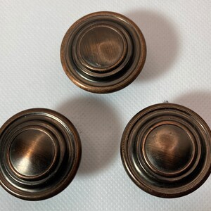 May include: Three bronze-colored cabinet knobs with a circular design. Each knob features concentric rings and a slightly domed center. The knobs have a brushed metal finish, giving them a vintage or antique appearance.