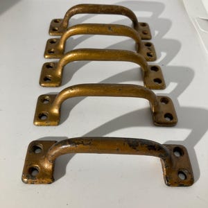 May include: Four vintage bronze-colored cabinet pulls. Each pull has a curved handle and a rectangular base with two screw holes. The pulls show signs of wear, with some areas of the bronze finish worn away.