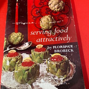May include: Vintage cookbook cover titled "serving food attractively" by Florence Brobeck. The cover features artichokes, a red candle, and a decorative metal piece. The book is set against a red background, with prepared artichokes filled with red caviar.