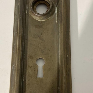 May include: A rectangular, antique brass door plate with a keyhole and a circular opening for a doorknob. The plate has a worn, aged appearance, with visible signs of use and age. The plate measures approximately 6 inches in length.