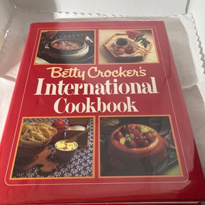 May include: A red hardcover cookbook titled "Betty Crocker's International Cookbook." The cover features four food images and the title in white lettering. The book is in a white box.