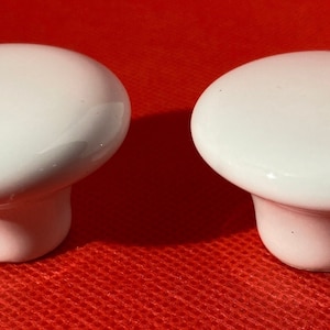 May include: Two white ceramic cabinet knobs with a glossy finish. The knobs have a rounded top and a short, cylindrical base. They are set against a red background, highlighting their clean, simple design. These knobs are suitable for furniture or cabinetry.