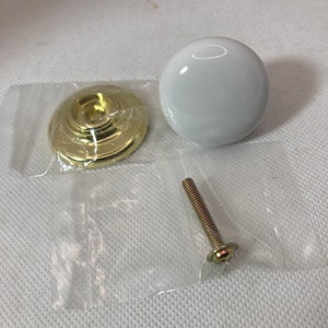 May include: A white, round cabinet knob with a gold-colored base and screw. The knob is glossy and appears to be ceramic. The hardware is packaged in a clear plastic bag, ready for installation.