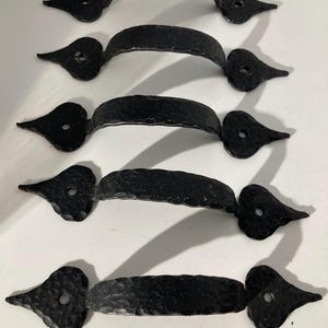May include: Five black, hammered-metal door pulls. Each pull has a curved handle and decorative, heart-shaped ends with a single hole for mounting. The pulls are arranged vertically against a white background.