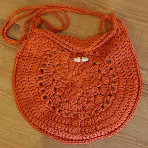 May include: A crocheted orange bag with a wooden button closure. The bag has a circular shape and a crocheted strap.