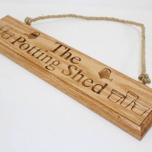 May include: A rectangular wooden sign with the words "The Potting Shed" engraved in a serif font. The sign features illustrations of flower pots and a watering can. A natural rope is attached for hanging.
