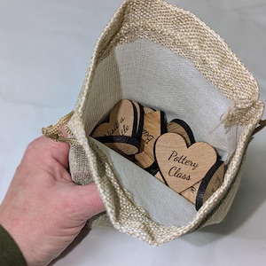 May include: A beige burlap drawstring bag containing wooden heart-shaped tokens. The tokens have black edges and engraved text, including "Pottery Class" and "Visit a little". The bag is held open, showing the contents.