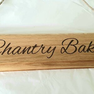 May include: Wooden sign with the text "Chantry Bakes" and rolling pin illustrations. The sign is made of light brown wood and has a rope for hanging. A decorative item for a kitchen or bakery.