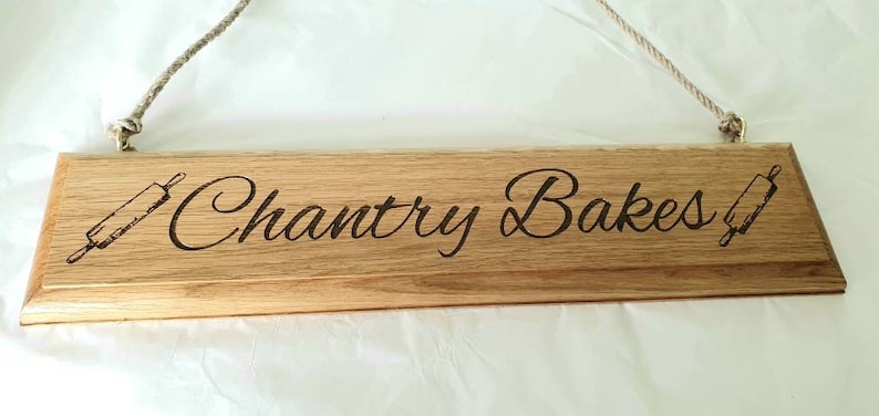 May include: Wooden sign with the text "Chantry Bakes" and rolling pin illustrations. The sign is made of light brown wood and has a rope for hanging. A decorative item for a kitchen or bakery.