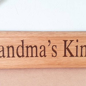 May include: Wooden sign with the words "Grandma's Kindy" engraved in a dark brown font. The sign has illustrations of children and is suspended by a beige rope. The sign is made of light brown wood.