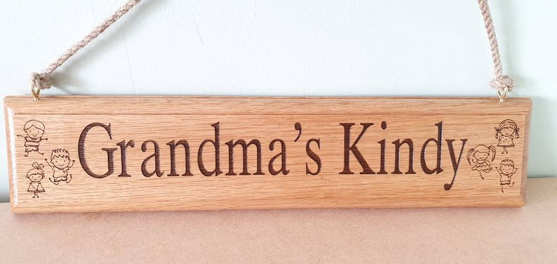 May include: Wooden sign with the words "Grandma's Kindy" engraved in a dark brown font. The sign has illustrations of children and is suspended by a beige rope. The sign is made of light brown wood.