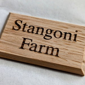 May include: A rectangular wooden sign with the engraved text "Stangoni Farm". The sign is made of light-coloured wood with visible grain and a bevelled edge. The text is in a classic serif font.