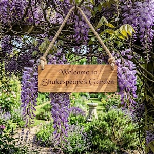 Può includere: Una targa rettangolare in legno marrone chiaro con venature naturali, recante il testo inciso "Welcome to Shakespeare's Garden" in un carattere serif scuro. La targa è appesa tramite una spessa corda ritorta di colore beige con occhielli.