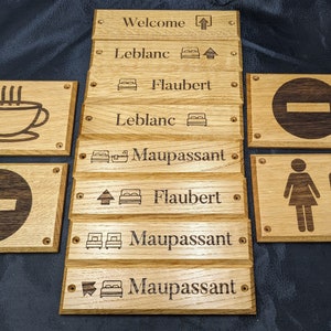 May include: A collection of rectangular light brown wooden signs with dark brown laser-engraved text and icons. The signs feature various labels including Welcome, Leblanc, Flaubert, and Maupassant, alongside symbols for beds, restrooms, and no-entry signs.