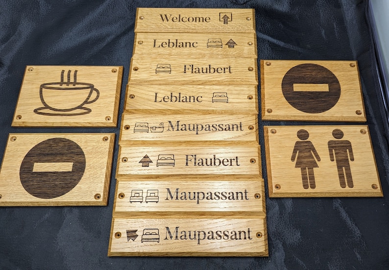 May include: A collection of rectangular light brown wooden signs with dark brown laser-engraved text and icons. The signs feature various labels including Welcome, Leblanc, Flaubert, and Maupassant, alongside symbols for beds, restrooms, and no-entry signs.