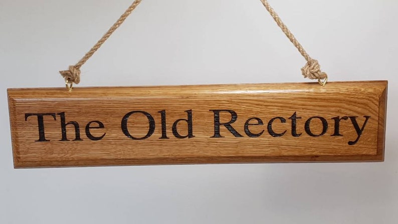 May include: Wooden sign with the text "The Old Rectory" engraved in a dark font. The sign is rectangular, made of light brown wood, and hangs from a natural rope. The sign is a decorative item.