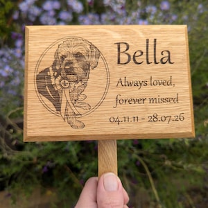 May include: A rectangular light brown wooden memorial plaque featuring a laser-engraved portrait of a dog wearing a ribbon, the name Bella, the text Always loved, forever missed, and the dates 04.11.11 - 28.07.26 on a wooden stake.