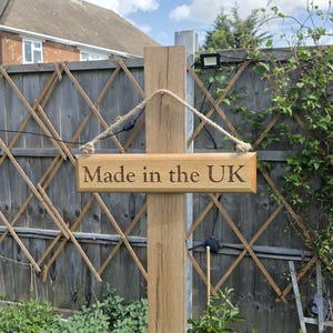May include: A wooden sign with the text "Made in the UK" hangs from a wooden post. The sign is suspended by a rope. The background includes a wooden fence and greenery.
