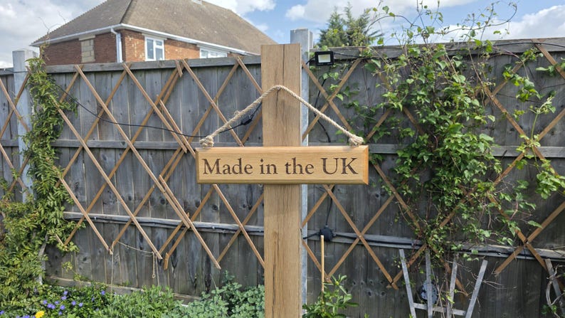 May include: A wooden sign with the text "Made in the UK" hangs from a wooden post. The sign is suspended by a rope. The background includes a wooden fence and greenery.