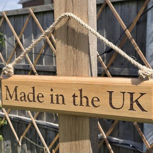 May include: Wooden sign with the text "Made in the UK" engraved in a serif font. The sign is suspended by a rope and mounted on a wooden post. The background includes a wooden fence and greenery.
