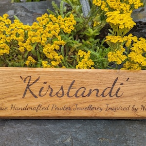 May include: A rectangular wooden sign with a natural grain finish featuring the engraved text 'Kirstandi' in a large script font, followed by the smaller engraved text 'Unique Handcrafted Pewter Jewellerey Inspired by Nature' below it.