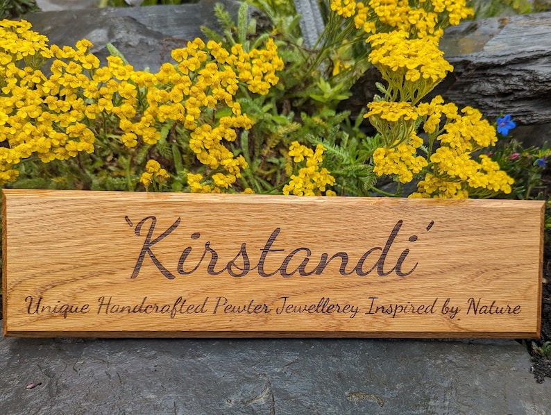 May include: A rectangular wooden sign with a natural grain finish featuring the engraved text 'Kirstandi' in a large script font, followed by the smaller engraved text 'Unique Handcrafted Pewter Jewellerey Inspired by Nature' below it.