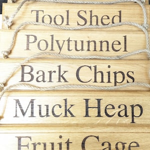 May include: A stack of wooden garden signs with dark brown engraved text. The signs read "Compost Heap", "Tool Shed", "Polytunnel", "Bark Chips", "Muck Heap", and "Fruit Cage". Each sign has a jute rope for hanging.