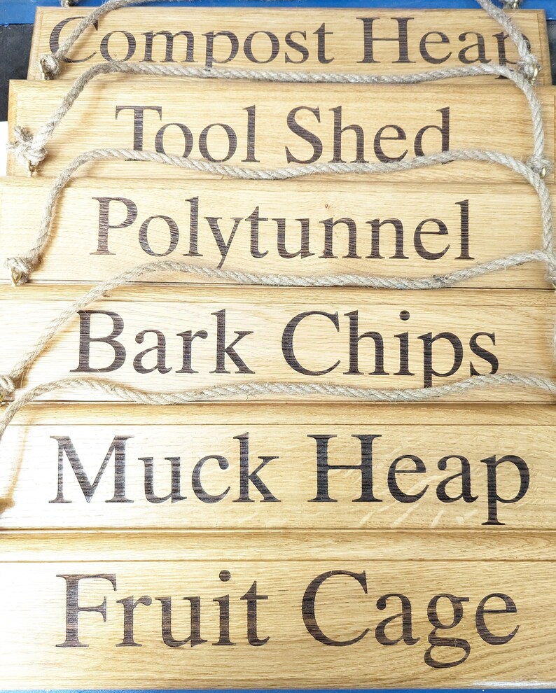 May include: A stack of wooden garden signs with dark brown engraved text. The signs read "Compost Heap", "Tool Shed", "Polytunnel", "Bark Chips", "Muck Heap", and "Fruit Cage". Each sign has a jute rope for hanging.