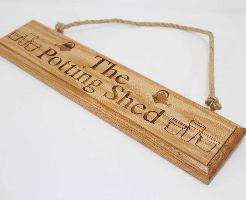 May include: A rectangular wooden sign with the words "The Potting Shed" engraved in a serif font. The sign features illustrations of flower pots and a watering can. A natural rope is attached for hanging.
