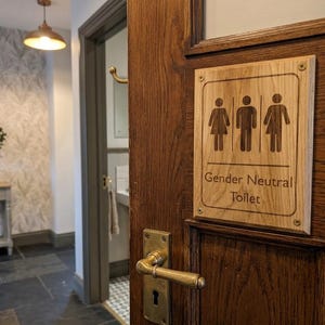 May include: A rectangular light-colored wooden sign mounted on a dark wood door with a brass handle. The sign features engraved icons of a person in a dress, a person in pants, and a person in a split garment, with the text Gender Neutral Toilet.