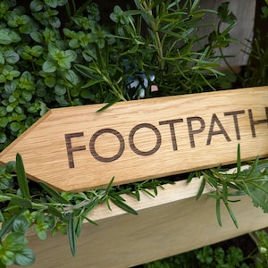 Puede incluir: Señal de madera en forma de flecha con la palabra "FOOTPATH" grabada en una fuente marrón oscuro. La señal está rodeada de hierbas y plantas verdes, lo que sugiere un jardín o un entorno al aire libre. La señal es de madera marrón claro.