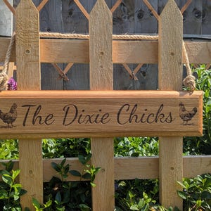 May include: Wooden sign with the text "The Dixie Chicks" and two engraved chicken illustrations. The sign is suspended by rope and mounted on a wooden fence. The sign is made of light brown wood.