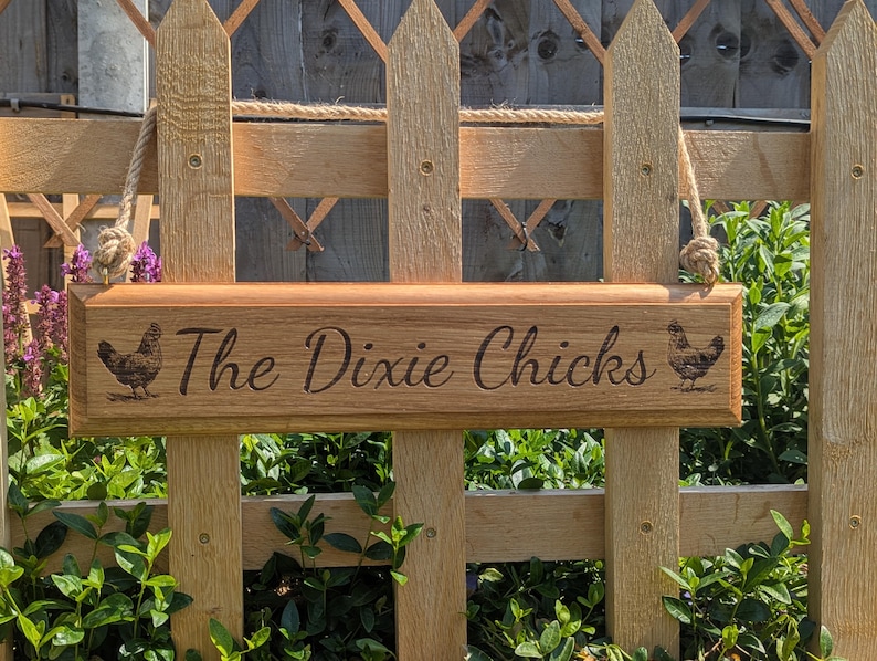 May include: Wooden sign with the text "The Dixie Chicks" and two engraved chicken illustrations. The sign is suspended by rope and mounted on a wooden fence. The sign is made of light brown wood.