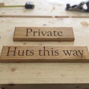 May include: Two rectangular light brown wooden signs with beveled edges. The top sign features the word "Private" engraved in a dark serif font. The bottom sign features the text "Huts this way" engraved in the same dark serif font.