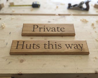 Puede incluir: Dos letreros rectangulares de madera marrón claro con bordes biselados. El letrero superior presenta la palabra "Private" grabada en una fuente serif oscura. El letrero inferior presenta el texto "Huts this way" en la misma fuente.